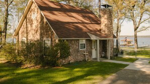 A stone house with a brown roof, a porch, and a view of a body of water. - Sunrise Shores - Waterfront Cottage sleeps 8.   (Chandler)