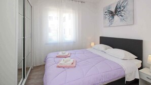 A bedroom with a bed, a window with sheer curtains, a wall-mounted artwork, and a bedside table with a lamp. - Holiday apartment with air conditioning near the beach (Baška)