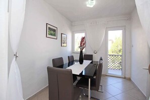 Un comedor con una mesa, sillas y un florero. Hay dos ventanas con cortinas, y una puerta que da a un espacio exterior.