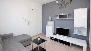 A modern living room with a grey sofa, a glass coffee table, a flat-screen TV, white shelving units, and a door with a handle. - Holiday apartment with air conditioning near the beach (Baška)