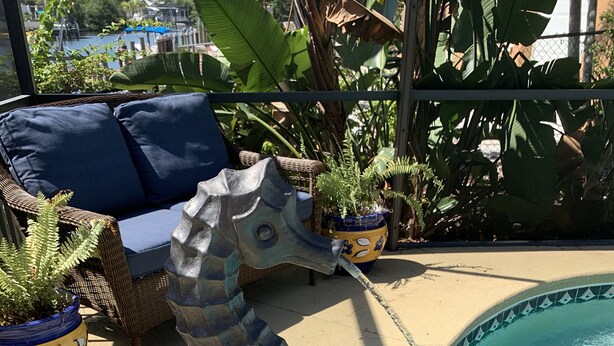 Een zeepaardbeeld langs een zwembad, met een rieten stoel en plantenpotten op de achtergrond.