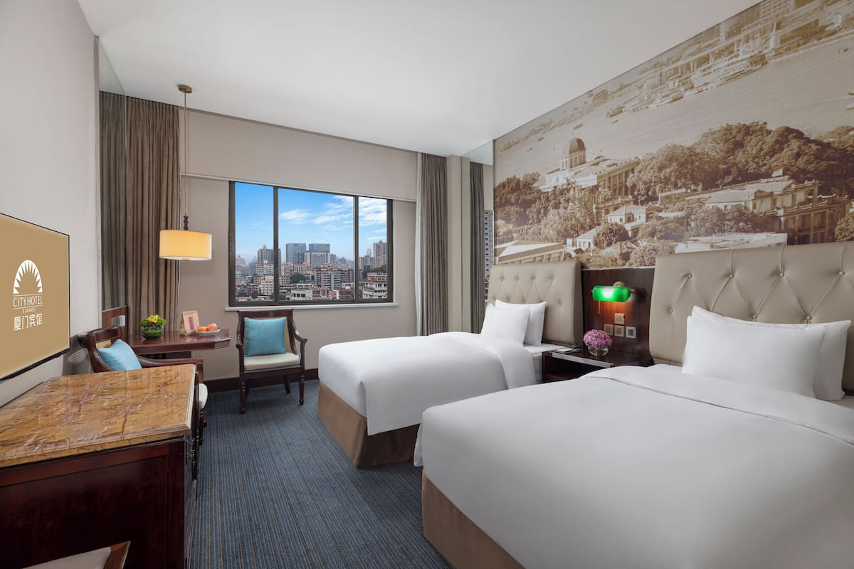A hotel room with two beds, a desk, a chair, a city view, and a large mural on the wall.