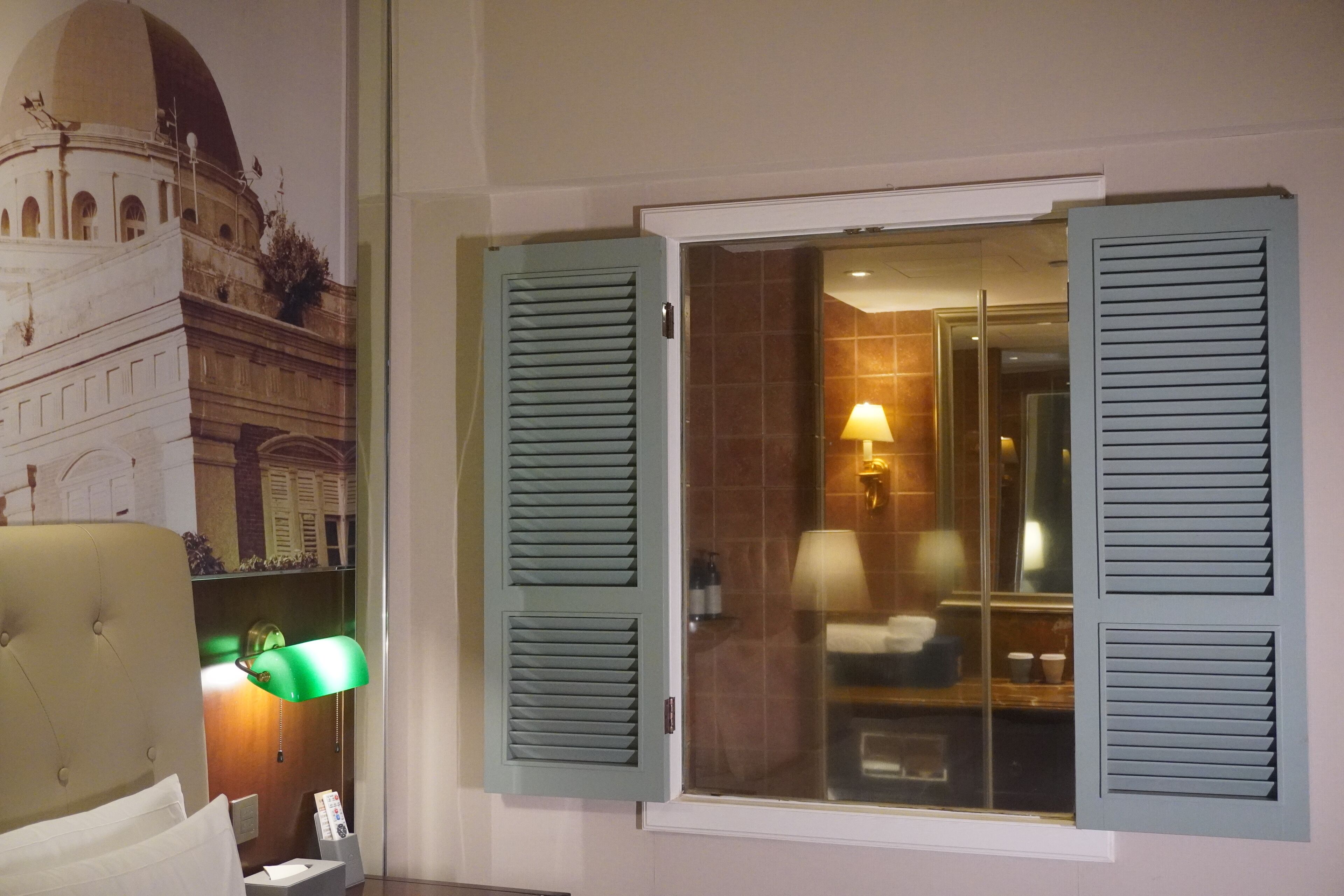 Una habitación de hotel con un ventanal grande, una cama con cabecera, una lámpara verde y un teléfono instalado en la pared.