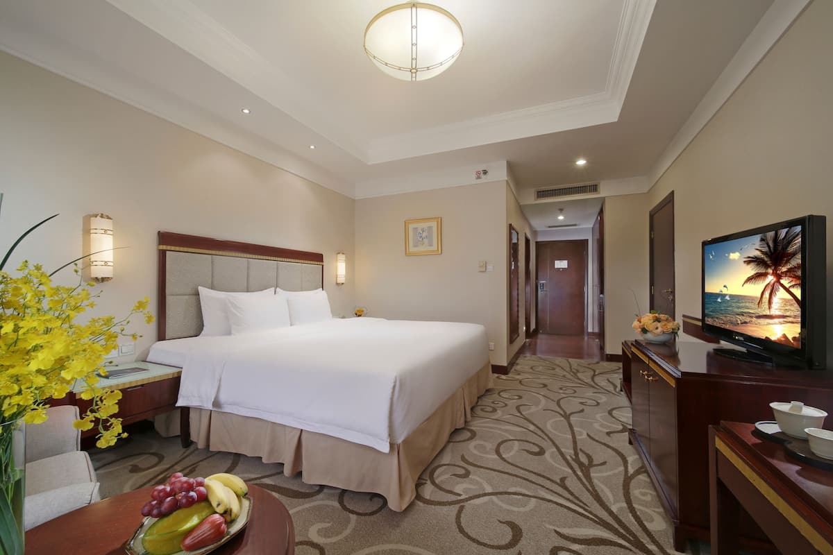 A hotel room with a large bed, a television, a desk with a bowl of fruit, and a vase of flowers.