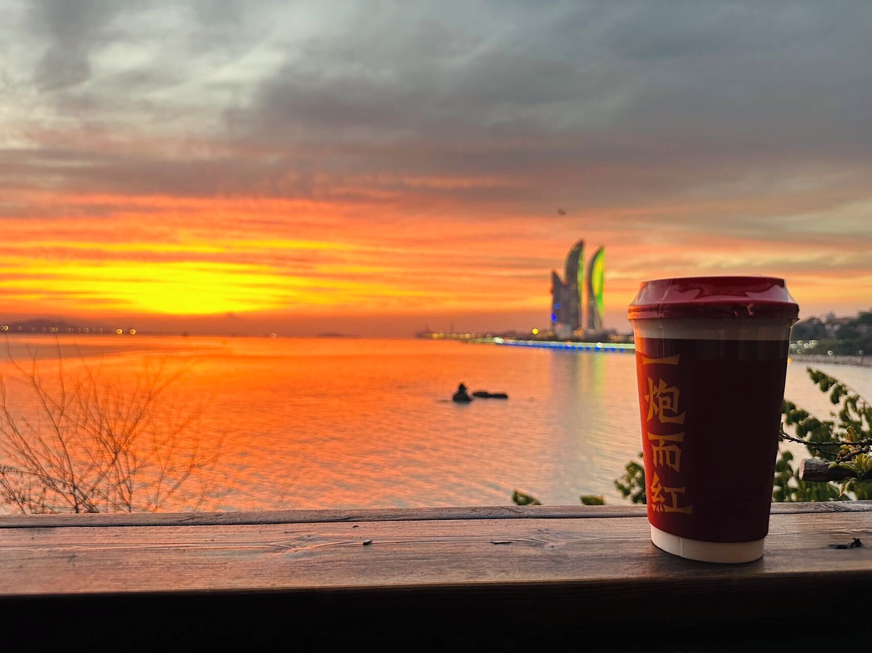 A wooden railing with a disposable cup in the foreground and a sunset over a body of water with a distant building.
