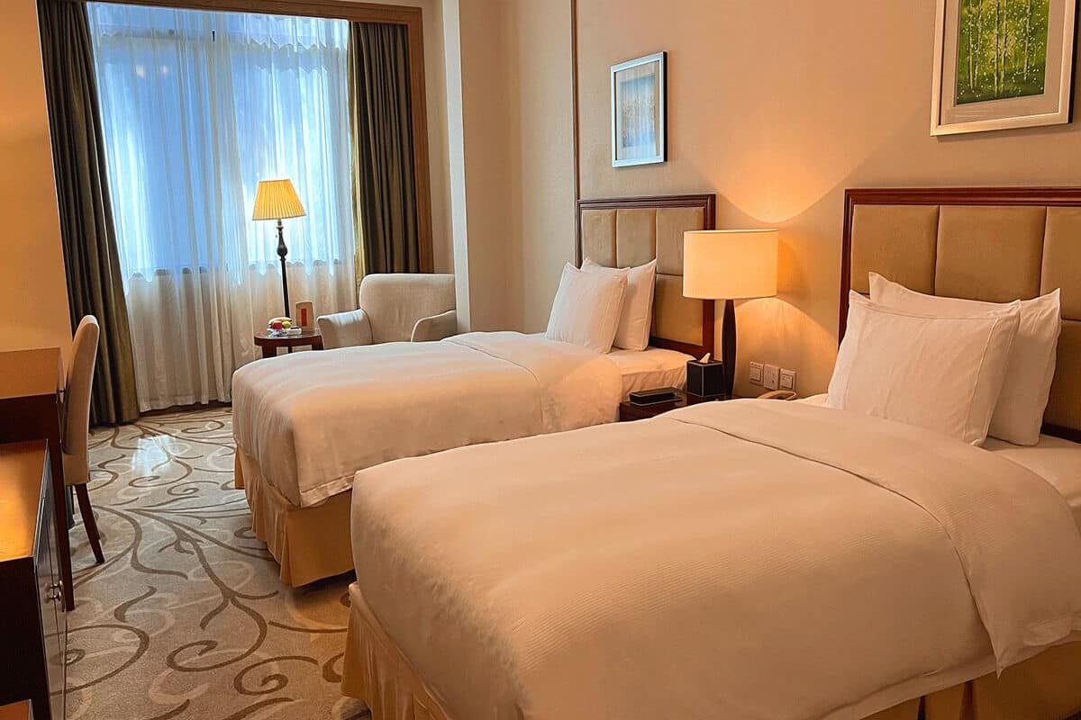 A hotel room with two beds, a desk, a chair, a lamp, and a window with curtains.