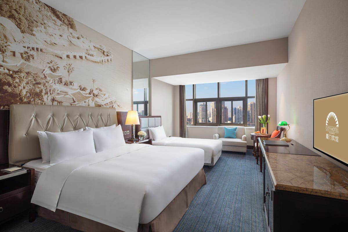 A hotel room with two beds, a sofa, a desk, and a large window offering a city view.