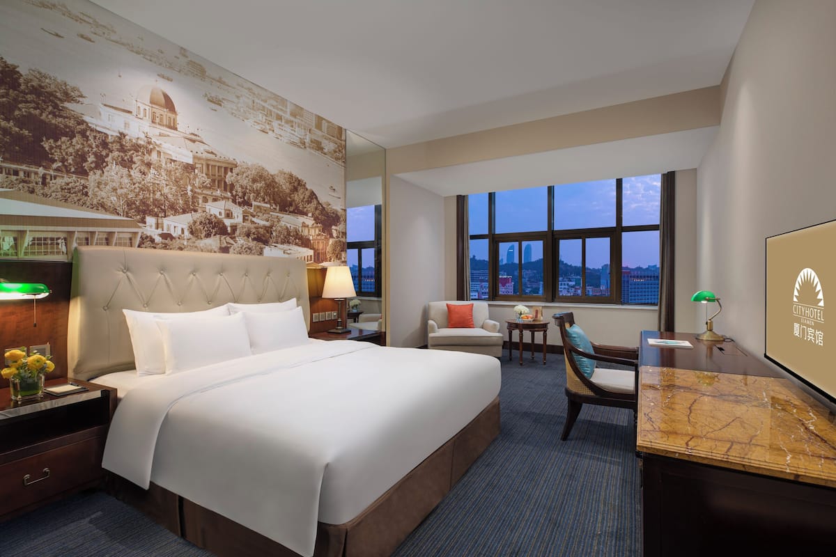 A hotel room with a large bed, a desk, a chair, and a view of the cityscape through large windows.