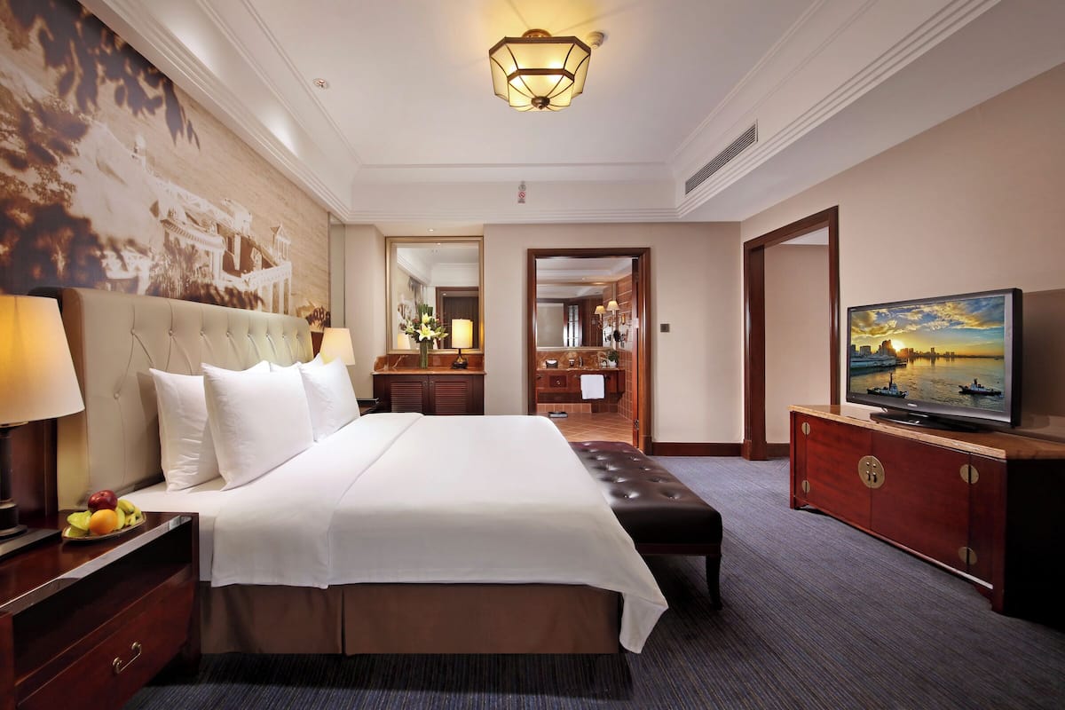 A hotel room with a large bed, a television, a desk with a lamp, and a wall with a cityscape painting.