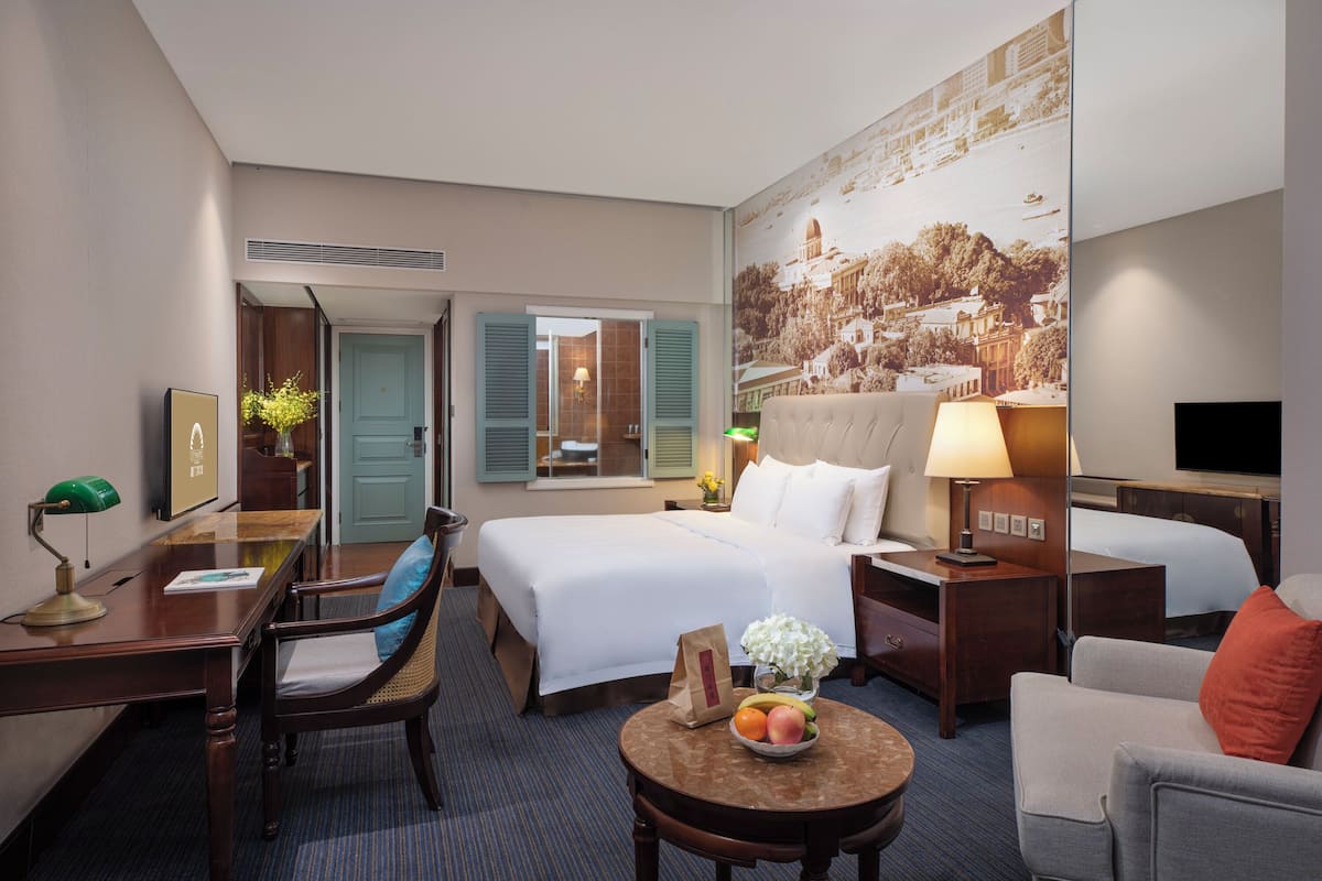 A hotel room with a large bed, a desk with a lamp, a chair, a small table with fruit, and a television.