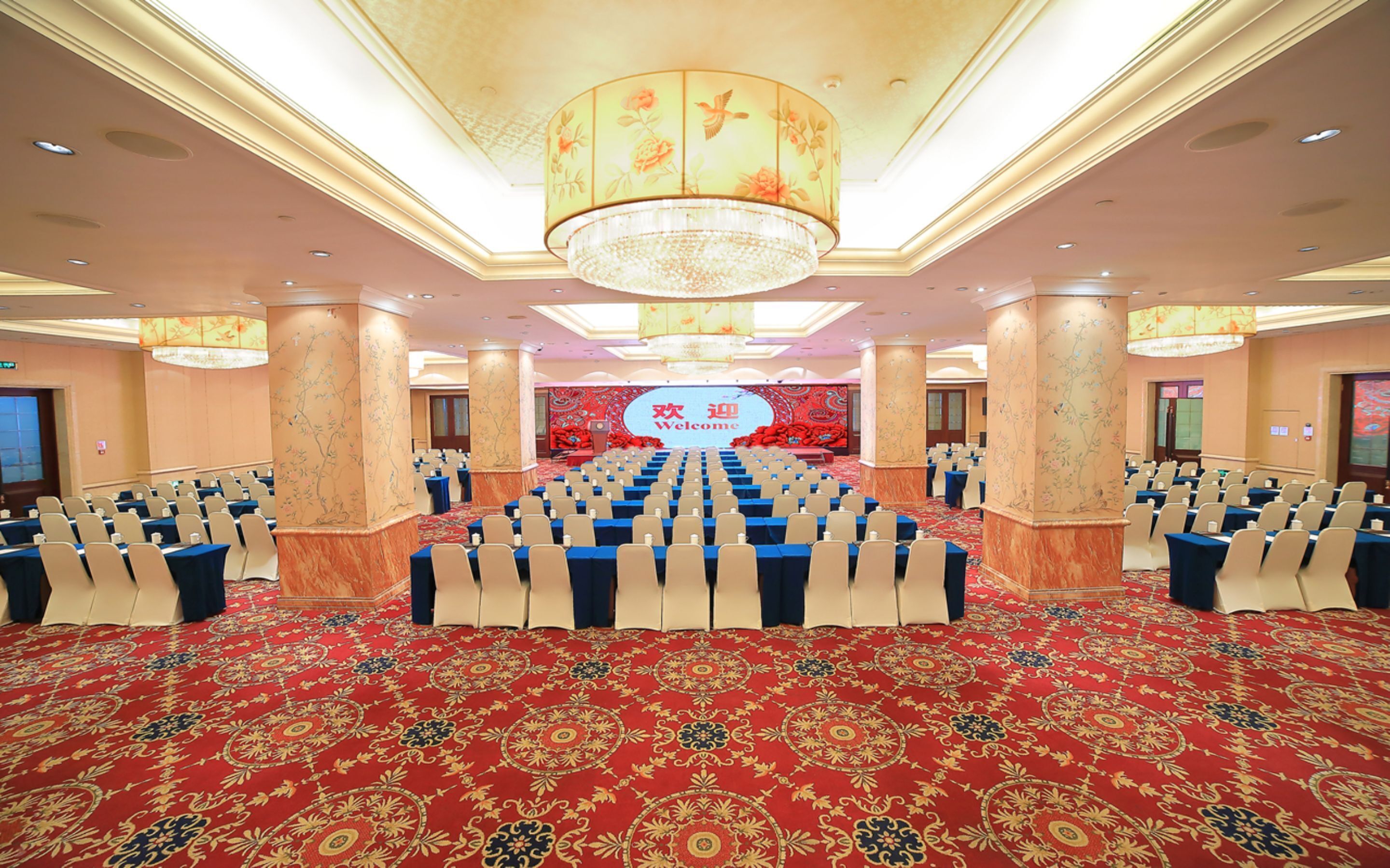A conference room with rows of chairs, a stage with a 'Welcome' sign, and a chandelier.