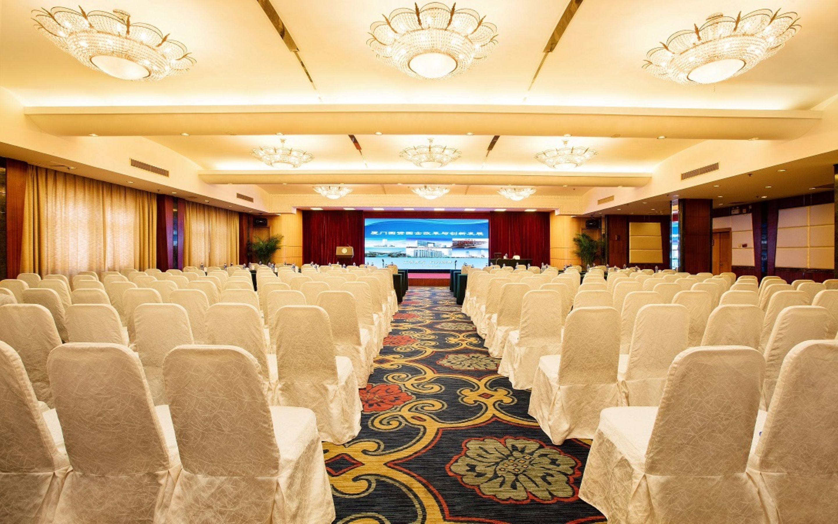 Un salón de conferencias con hileras de sillas, piso alfombrado y candelabros.
