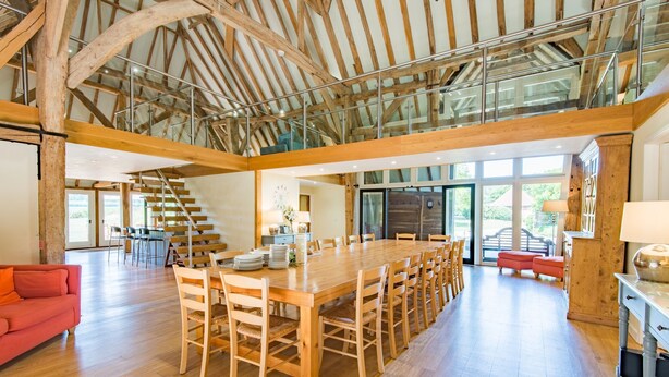 A spacious dining area with a long wooden table and chairs, a red sofa, and a staircase leading to an upper level. - Great Higham Barn by Bloom Stays -  sleeps 26 guests  in 7 bedrooms (Sittingbourne)