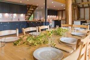 A dining table set with plates, glasses, and cutlery, surrounded by chairs in a modern kitchen with dark cabinets and wooden beams. - Great Higham Barn by Bloom Stays -  sleeps 26 guests  in 7 bedrooms (Sittingbourne)