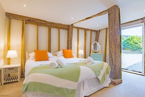 A bedroom with a large bed, wooden beams, a nightstand, a dresser, and a window with a view of greenery. - Great Higham Barn by Bloom Stays -  sleeps 26 guests  in 7 bedrooms (Sittingbourne)