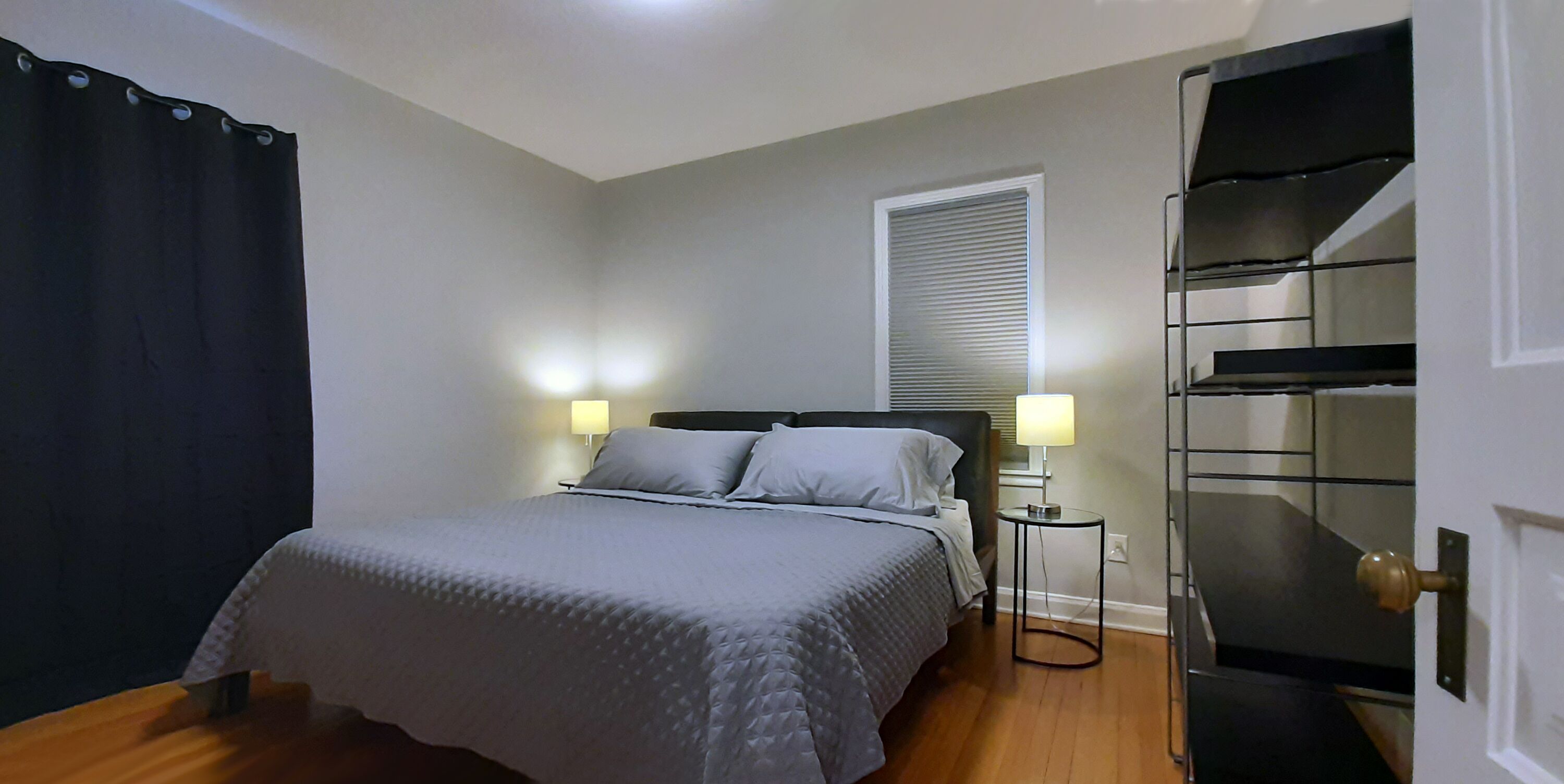 Une chambre à coucher avec un lit, des lampes de chevet, une fenêtre avec des stores, une armoire noire et une porte dotée d’une poignée en laiton.