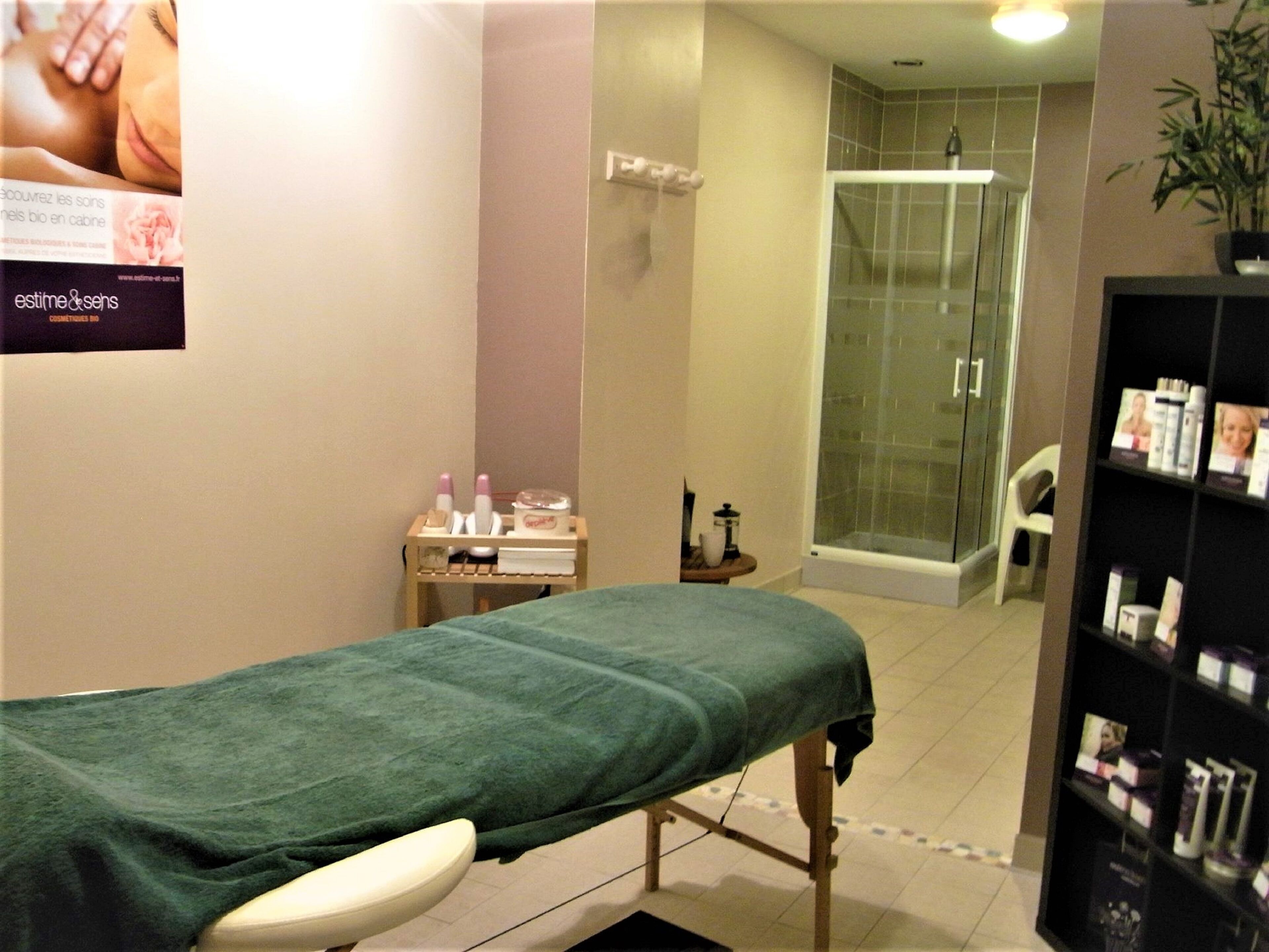 Une salle de soins équipée d’une table de massage, d’une petite étagère avec des flacons, d’une cabine de douche en verre et d’une autre étagère contenant des produits.