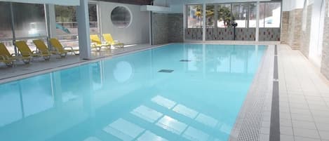 An indoor swimming pool with clear water, yellow lounge chairs, and a high ceiling with wooden beams.