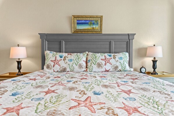 Una cama con colcha estampada de flores y estrellas de mar, dos almohadas a juego, un cuadro enmarcado sobre la cabecera y dos lámparas de noche sobre mesitas de madera.