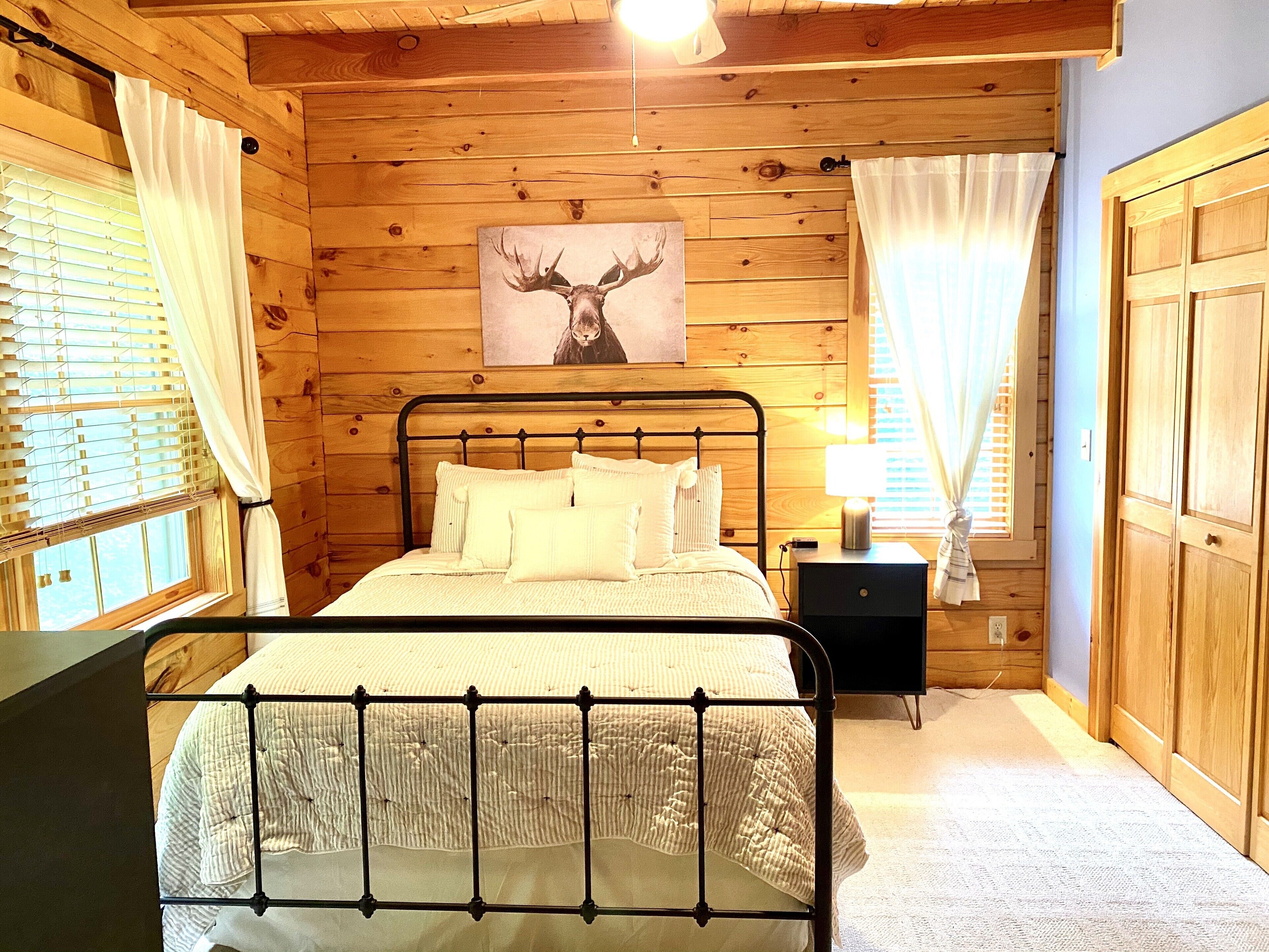 A wooden cabin bedroom with a bed, a nightstand, a lamp, and a painting of a moose's head on the wall.