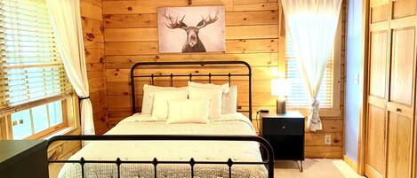 A wooden cabin bedroom with a bed, a nightstand, a lamp, and a painting of a moose's head on the wall.