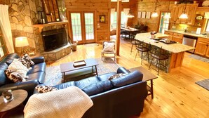 A spacious living area with a stone fireplace, a black leather sofa, a dining table, and a kitchen with wooden cabinets and a bar area.