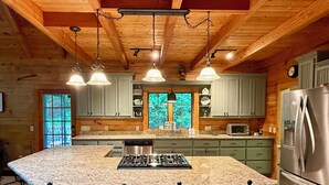A kitchen with granite countertops, a central island with bar stools, wooden ceiling beams, and a stainless steel refrigerator.