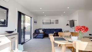 Ein modernes Wohnzimmer mit einer Couch, einem Esstisch und Stühlen. Ein Fernseher ist an der Wand befestigt und ein Fenster mit Jalousien vorhanden.
