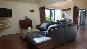 A spacious living room with a large black leather sofa, a flat-screen TV mounted on the wall, and a dining area with a table and chairs. - Ahi Apartment  with Tasman Sea views (Raglan)