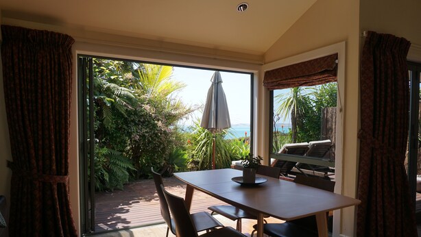 A dining area with a table and chairs, a potted plant, and a view of a garden and ocean through a glass door. - Ahi Apartment  with Tasman Sea views (Raglan)