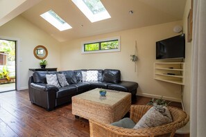 A living room with a black leather sofa, a wicker coffee table, a television on a stand, and a large window with a view of greenery.