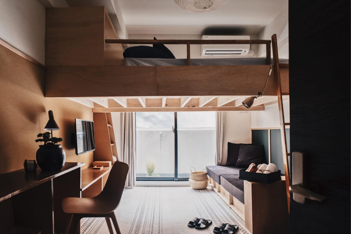 A compact, modern room with a bunk bed, a desk, a chair, a sofa, and a large window offering a view of the outdoors.