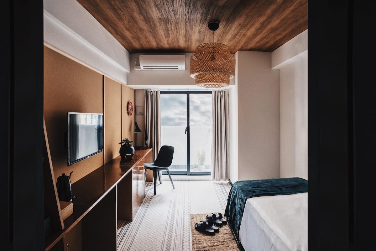 A modern hotel room with a wooden ceiling, a flat-screen TV, a desk, a chair, a bed, and a large window with curtains.