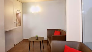 Ein Check-in-Bereich mit einer Rezeption, einem kleinen Tisch mit Pflanze und einer Sitzecke mit einer Couch und roten Kissen.