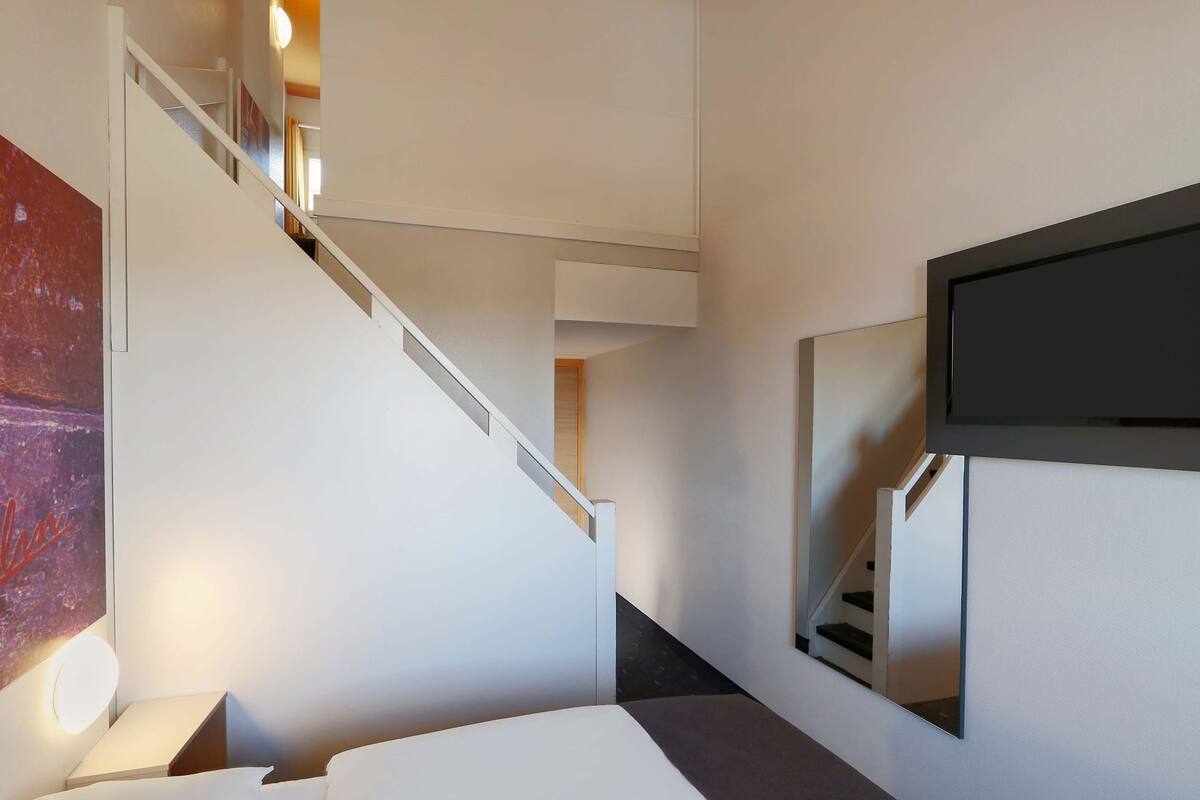 A modern hotel room with a bed, a TV mounted on the wall, and a staircase leading to another level.