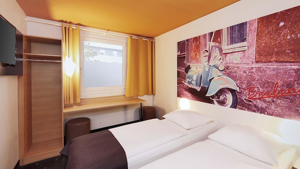 Ein Hotelzimmer mit zwei Betten, einem Kleiderschrank, einem Fenster mit Vorhängen und einem Wandbild eines Oldtimers.