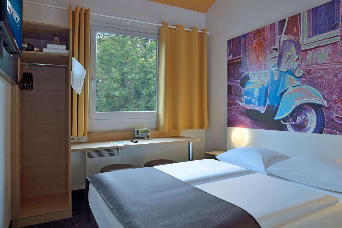 Une chambre d’hôtel équipée d’un lit, d’un bureau, d’une chaise, d’une armoire et d’une fenêtre avec des rideaux.