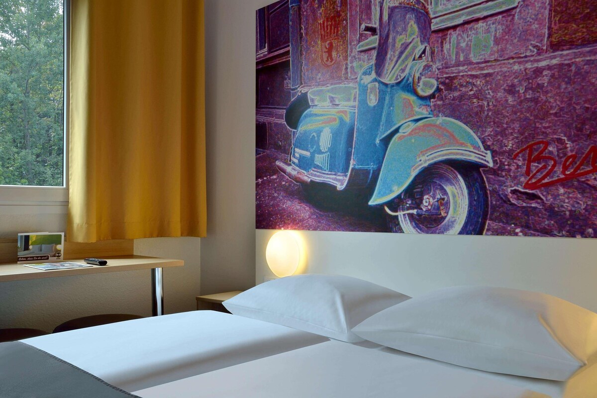 A hotel room with a bed, a desk with a lamp, and a wall with a vintage car painting.