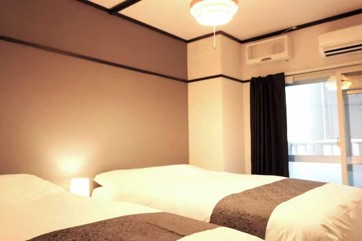A hotel room with two beds, a ceiling light, an air conditioning unit, and a window with curtains.