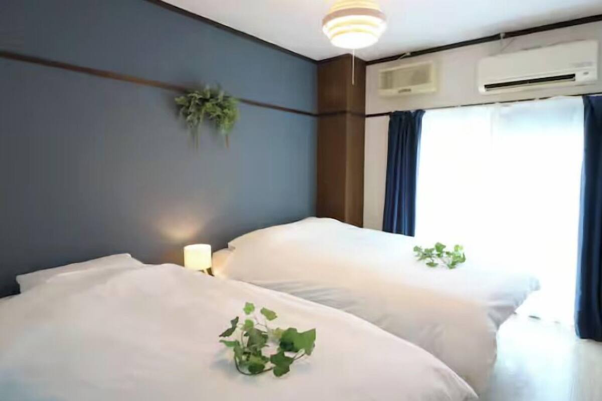 A hotel room with two beds, a wall-mounted air conditioner, a ceiling light fixture, and a window with curtains.