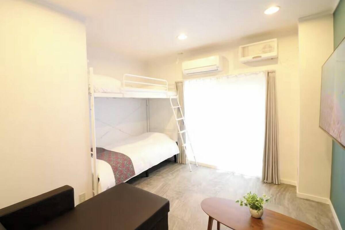 A compact hotel room with a bunk bed, a desk, a chair, a small table with a plant, and a window with curtains.