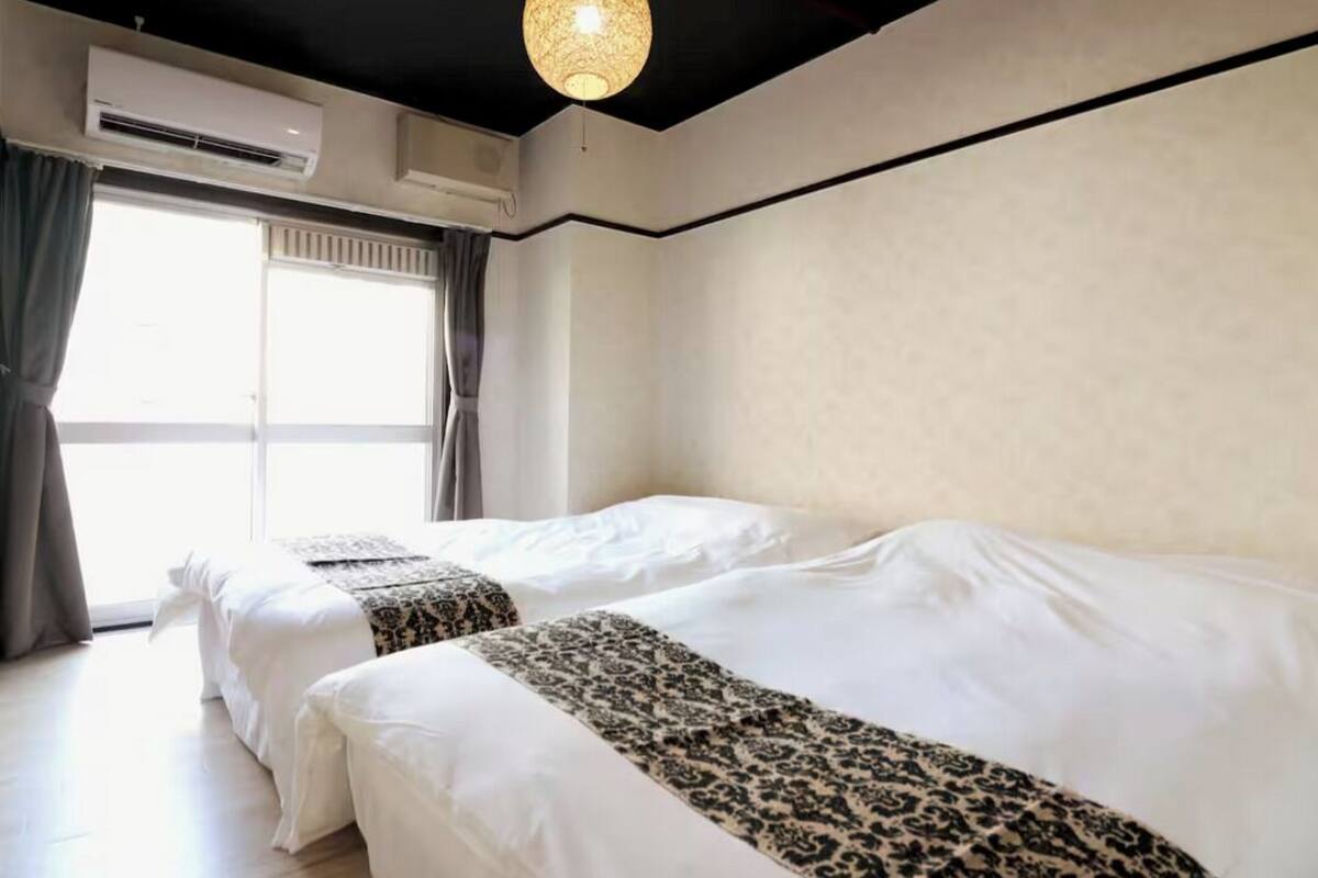 A hotel room with two beds, a large window with curtains, an air conditioning unit, and a hanging light fixture.
