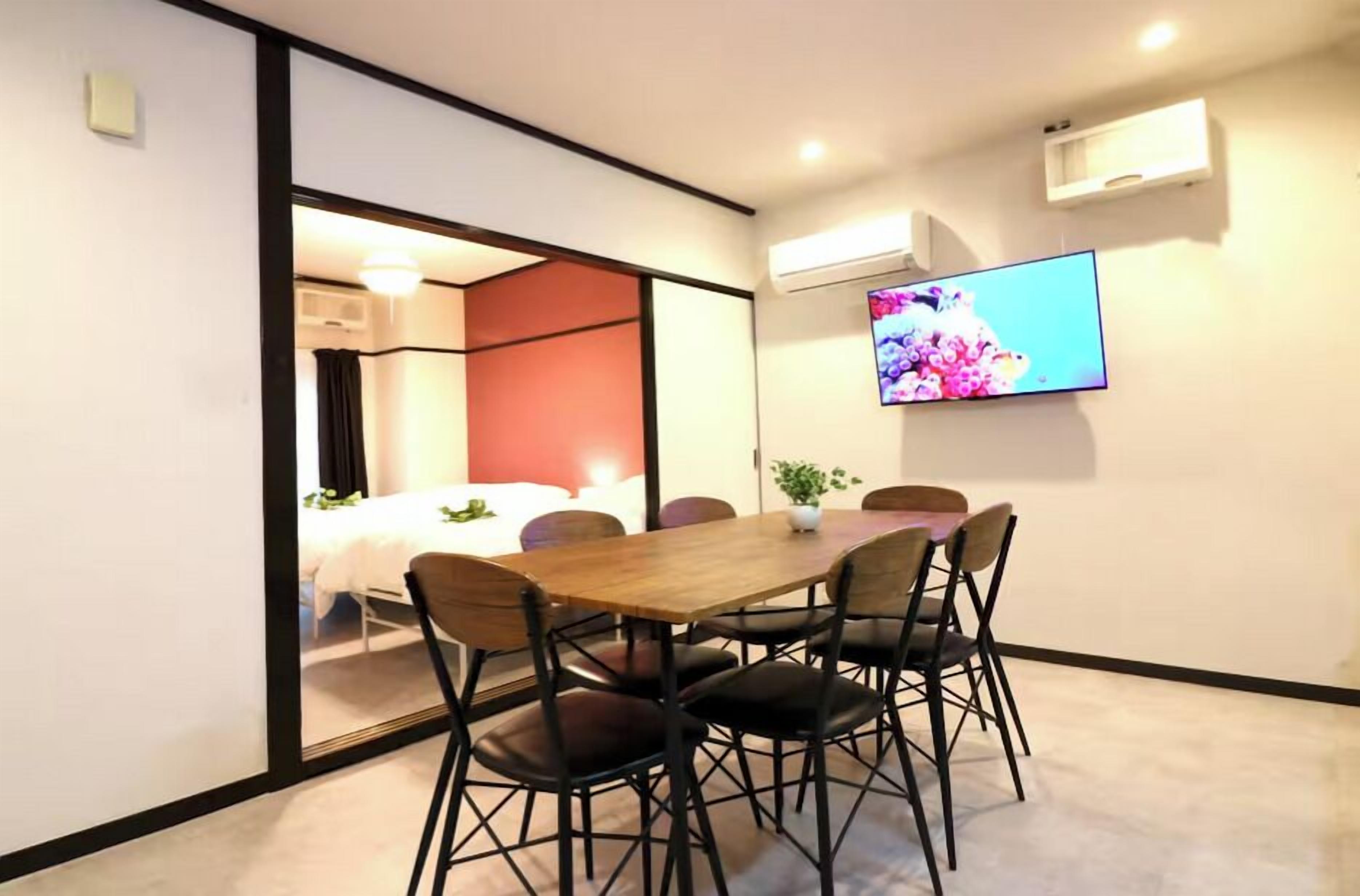 A conference room with a wooden table, black chairs, a TV on the wall, and a sliding door leading to a bedroom.