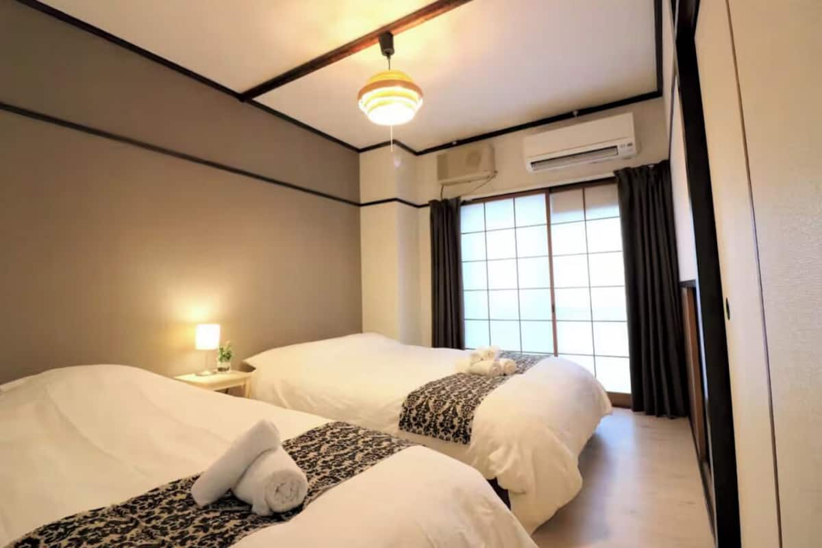 A hotel room with two beds, a bedside table with a lamp, an air conditioning unit, and a window with curtains.