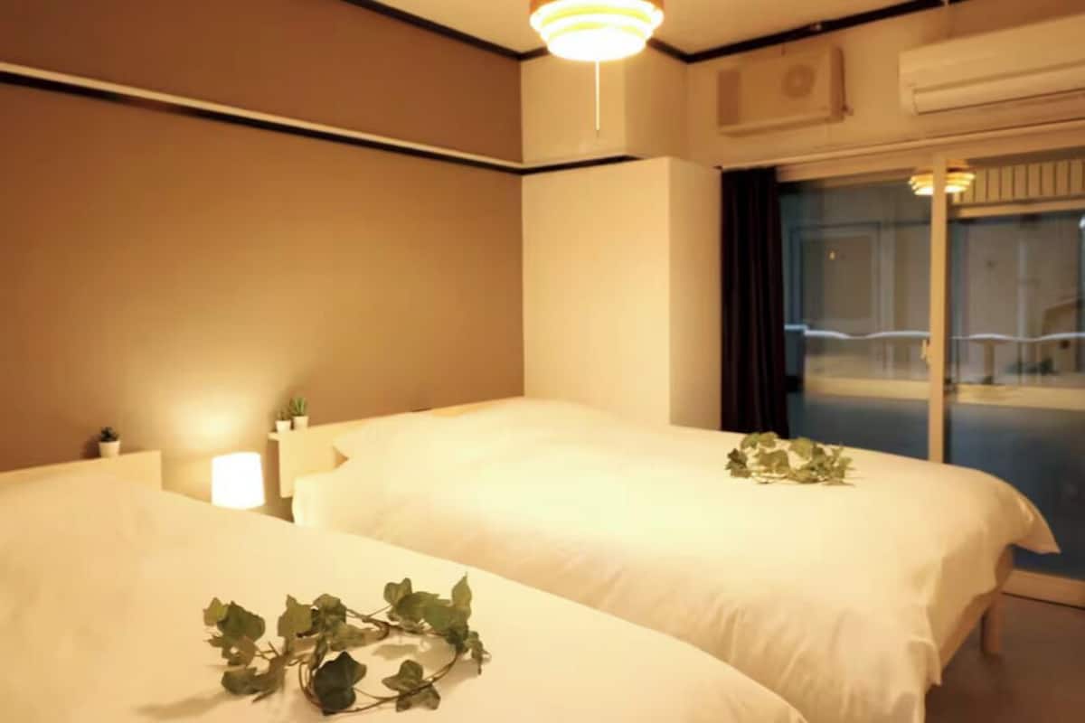 A hotel room with two beds, a bedside table with a lamp, and a window with a view of a balcony.