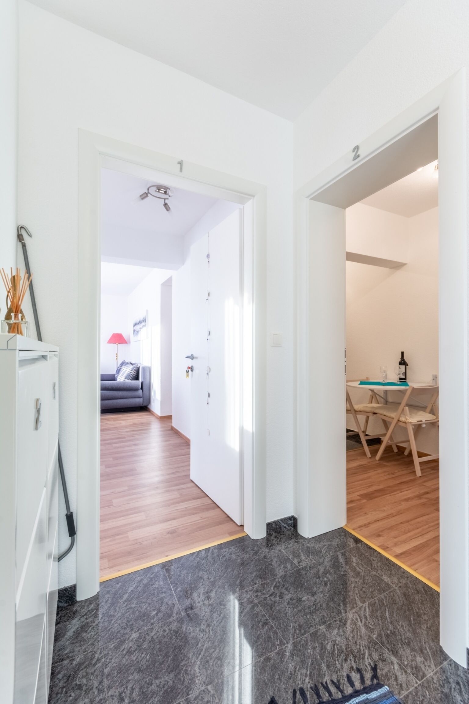 A hallway with a marble floor, a door leading to a room with a wooden floor, and a small table with a bottle.