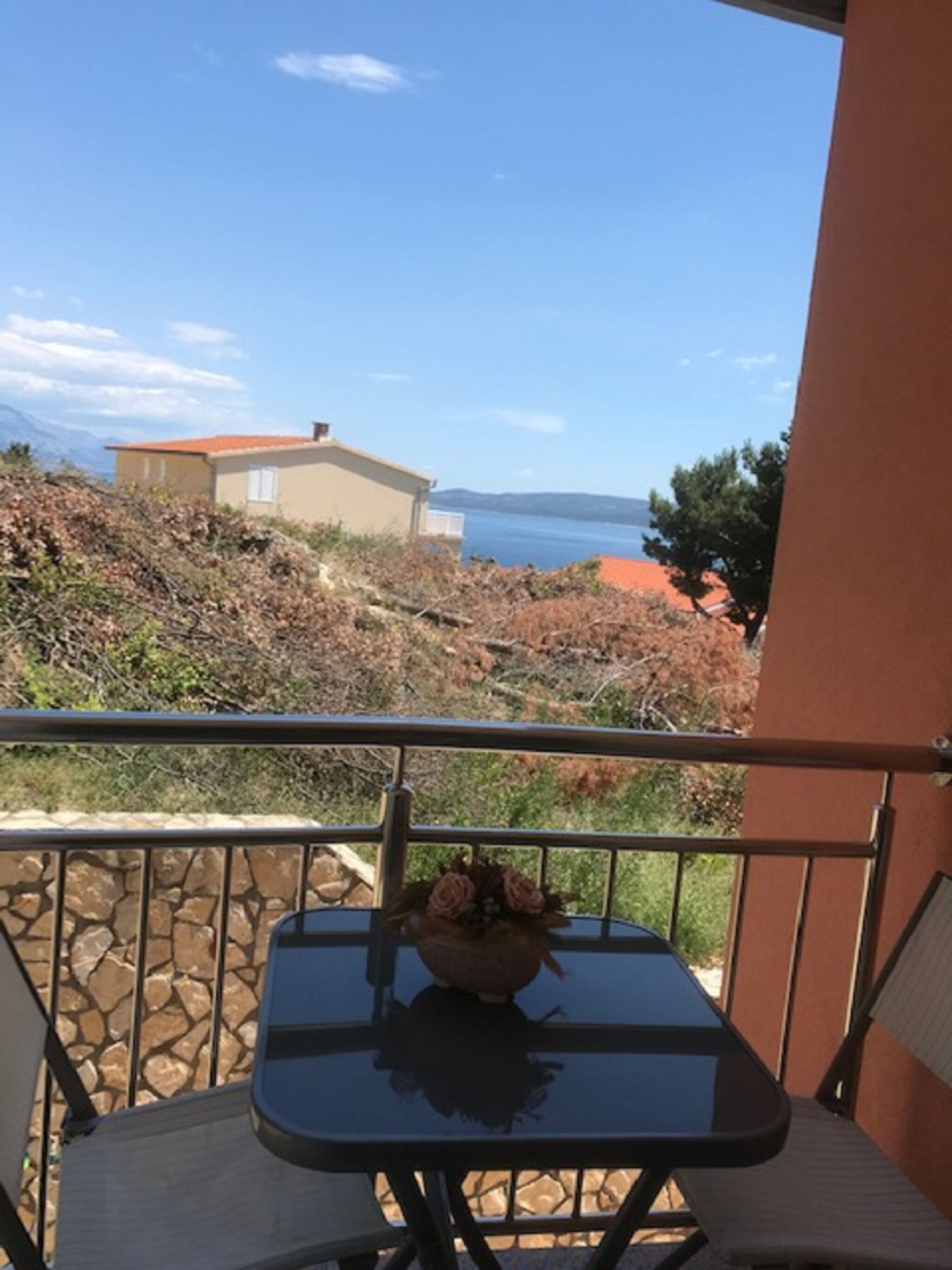 A balcony with a table and chairs, overlooking a coastal landscape with a house and a clear sky.