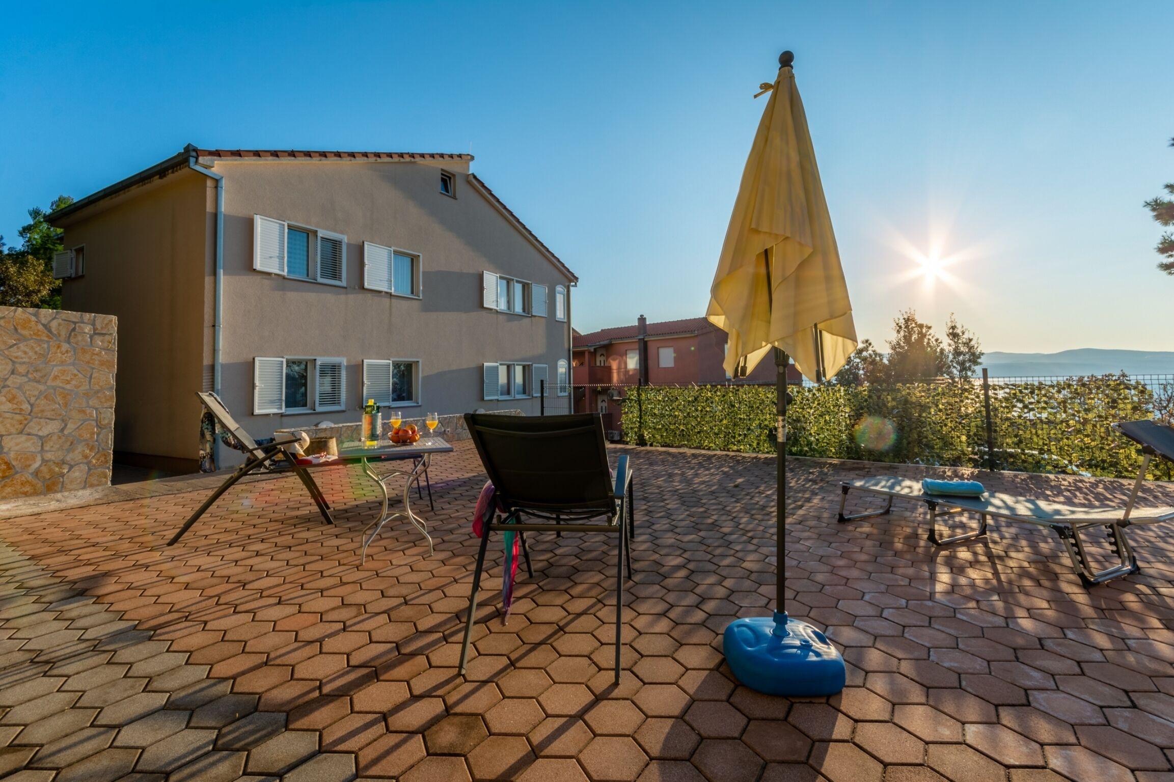 A patio with hexagonal tiles, a chair, a table with a bottle, and a yellow umbrella.