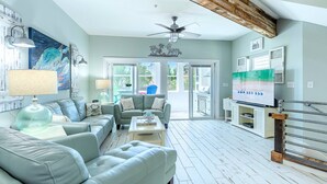 A spacious living room with a large sofa, a coffee table, and a television. There is a ceiling fan and a door leading to an outdoor area. - Modern Townhome, Short Walk to Beach w/ Private Access, Pool & Grill (Destin)