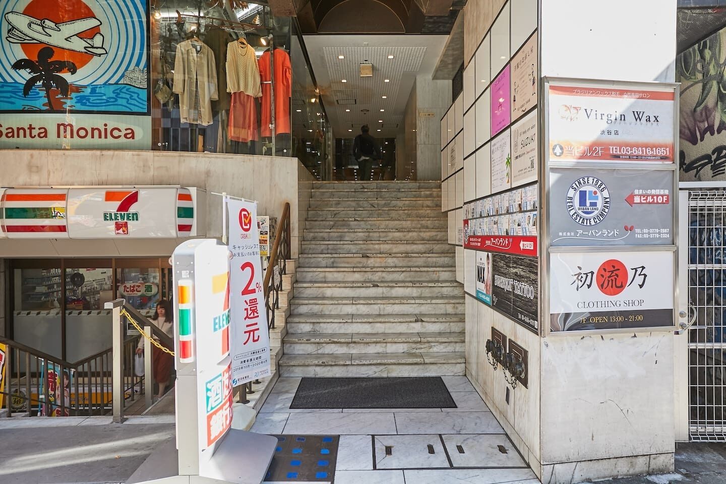 Una escalera conduce a un área comercial con varias tiendas, incluyendo una tienda de conveniencia 7-Eleven y una tienda de ropa.